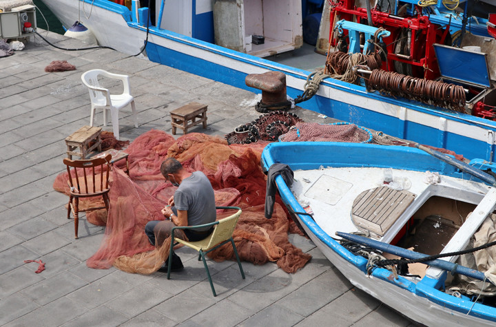 A local fisherman in Cetara sitting on the pier while mending traditional orange fishing nets next to a blue boat.