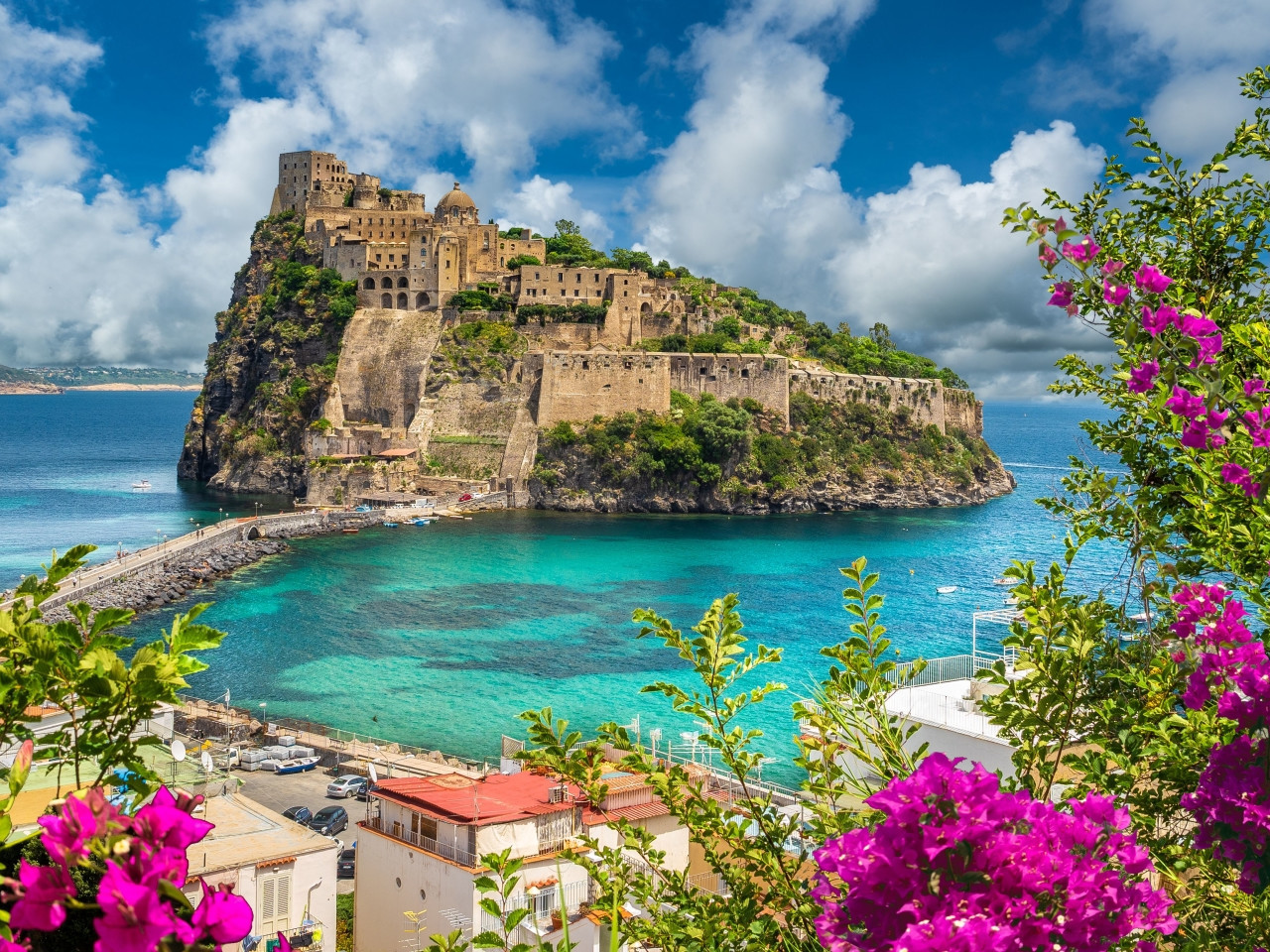 Majestic panoramic view of the Aragonese Castle in Ischia overlooking crystal-clear waters with pink bougainvillea flowers in the foreground.