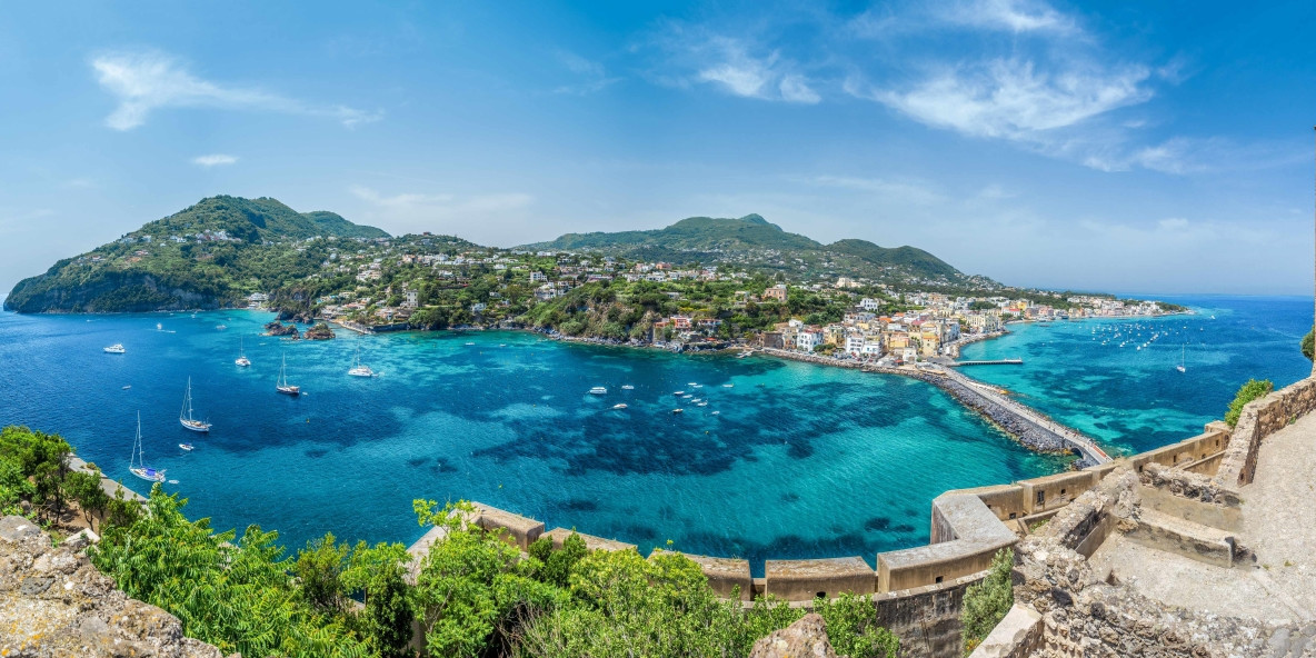 Wide panoramic view from the walls of the Aragonese Castle in Ischia, showing the turquoise bay waters, the connecting bridge, and the coastal village under a blue sky.