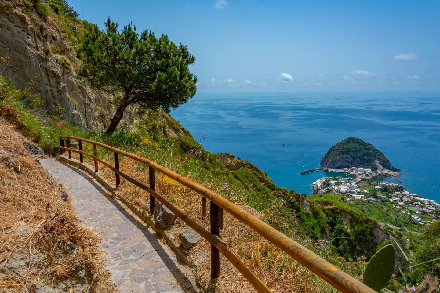 A paved hiking path with a wooden fence runs along a hillside in Ischia, offering a high-angle view of Sant'Angelo village and the deep blue sea.