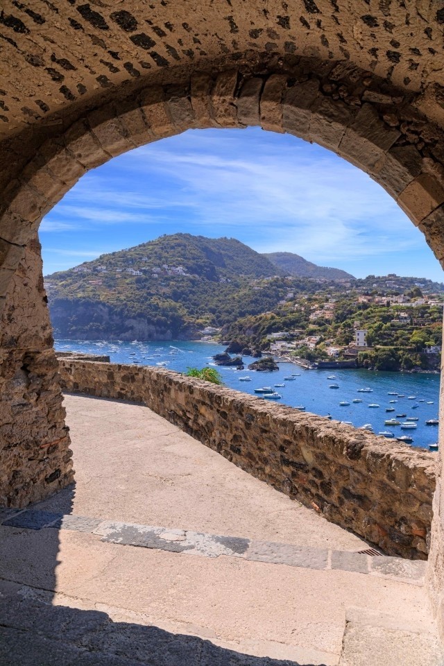 Scenic view of Cartaromana Bay in Ischia framed by a stone arch of the Aragonese Castle, with boats anchored in the open sea.