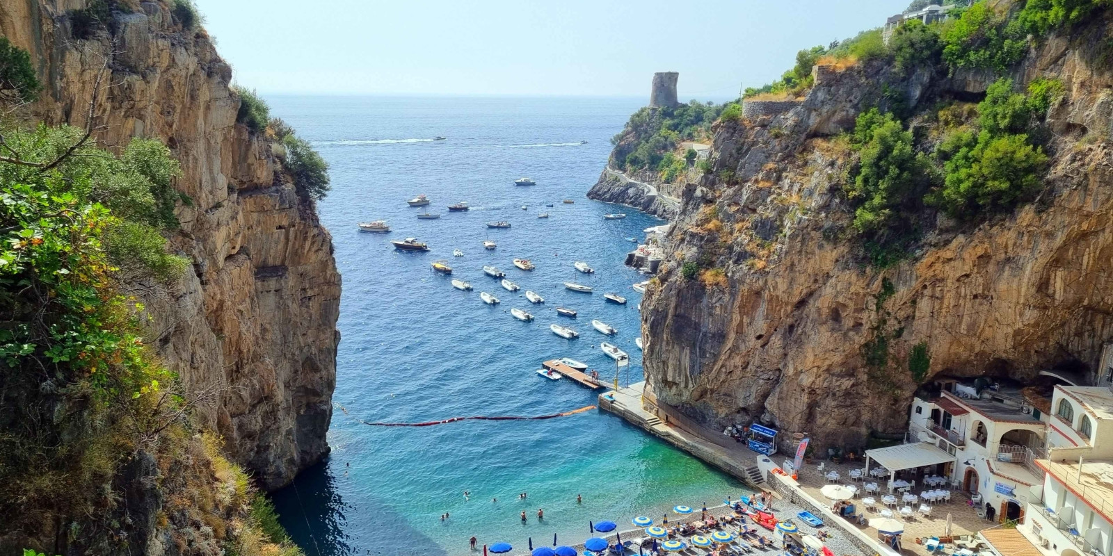 Stunning high-angle view of Marina di Praia bay in Praiano, featuring boats in crystal clear waters and a historic Saracen tower in the distance.