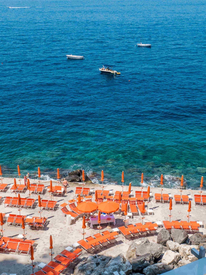 Stabilimento balneare a Praiano con file di ombrelloni arancioni su una piattaforma rocciosa affacciata sul mare turchese della Costiera Amalfitana.