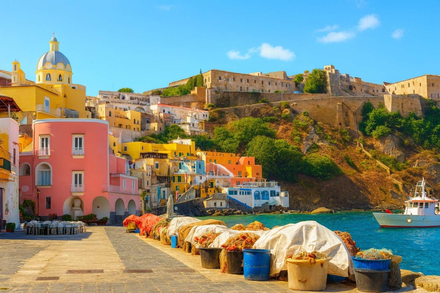 Il borgo di Marina Corricella a Procida visto dal molo, con reti da pesca in primo piano, case color pastello e l'abbazia di San Michele che sovrasta il porto.