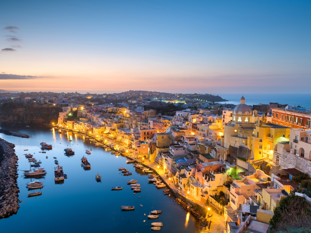 Vista aerea al tramonto di Marina Corricella a Procida con luci del borgo, case colorate e porto turistico.