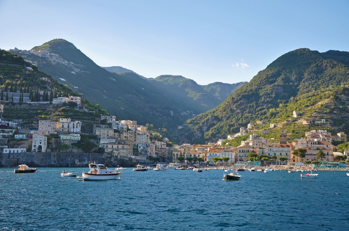 Scenic view of Maiori town from the water with boats anchored in the blue sea and mountains in the background.