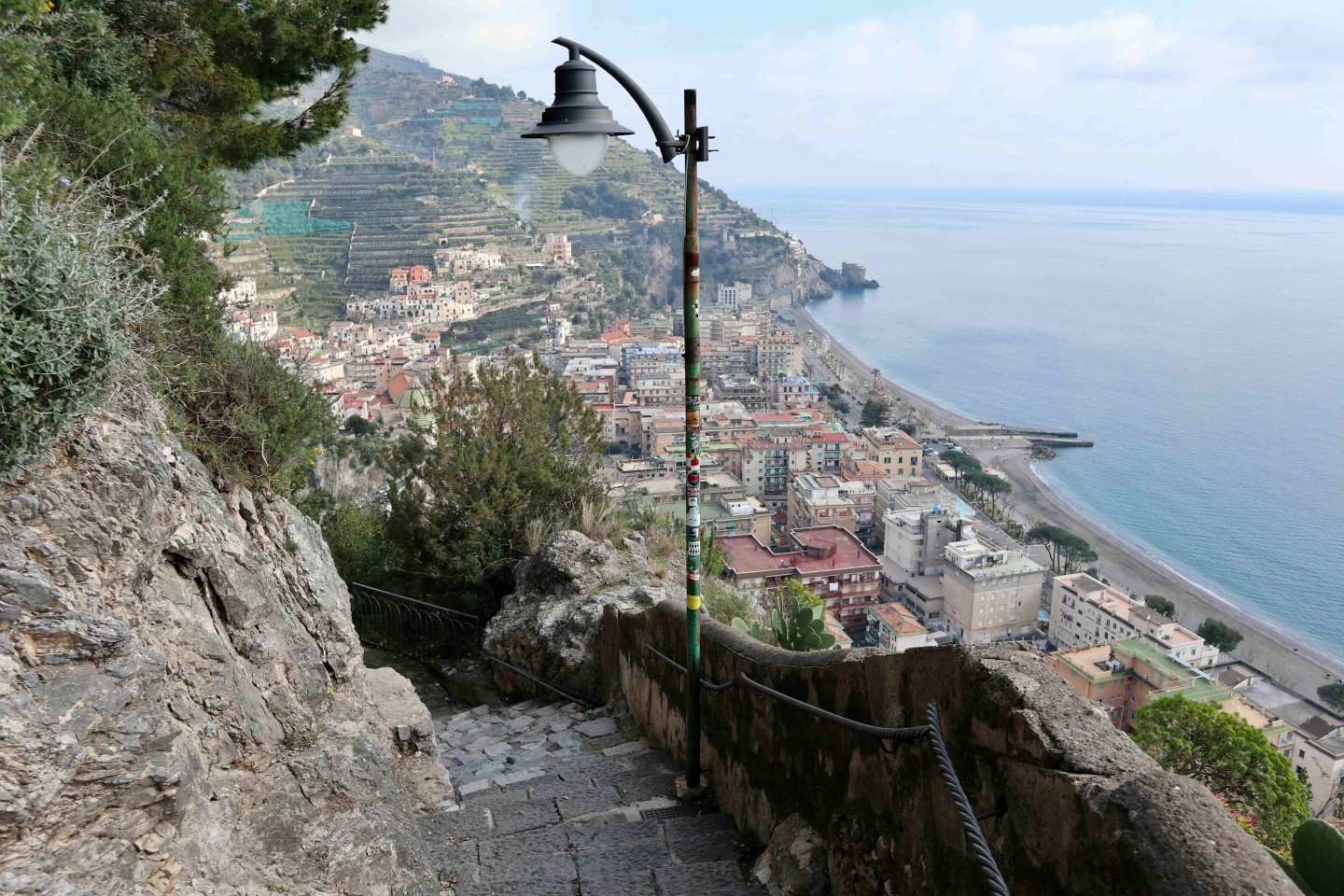 Panoramic view from a stone staircase overlooking Maiori town and beach on the Amalfi Coast with a vintage street lamp in the foreground.
